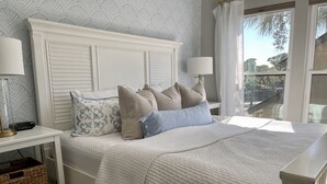 A neatly made bed with a white headboard, a bedside table with a lamp, and a view of the outdoors through a window. - Tennis Courts in front, and the ever changing Marsh in back. Golf cart included. (Fripp Island)