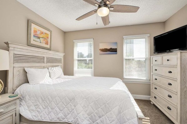 A bedroom with a bed, a ceiling fan, a dresser, a television, and two windows with blinds.