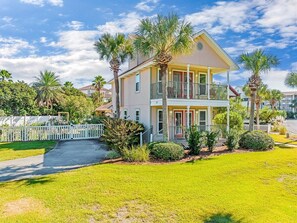 A two-story house with a balcony, surrounded by a white picket fence, palm trees, and a well-maintained lawn.