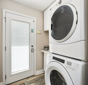 A laundry room with a front-loading washer and dryer stacked on top of each other, a door with a window, and a small shelf with a decorative item.