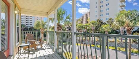A balcony with wooden chairs and a table, overlooking a street with palm trees and a multi-story building.
