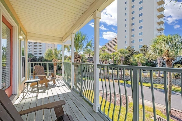 A balcony with wooden chairs and a table, overlooking a street with palm trees and a multi-story building.