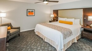 Une chambre d’hôtel avec un grand lit, deux tables de chevet avec des lampes, un ventilateur de plafond, un tableau encadré au mur et une moquette à motifs.