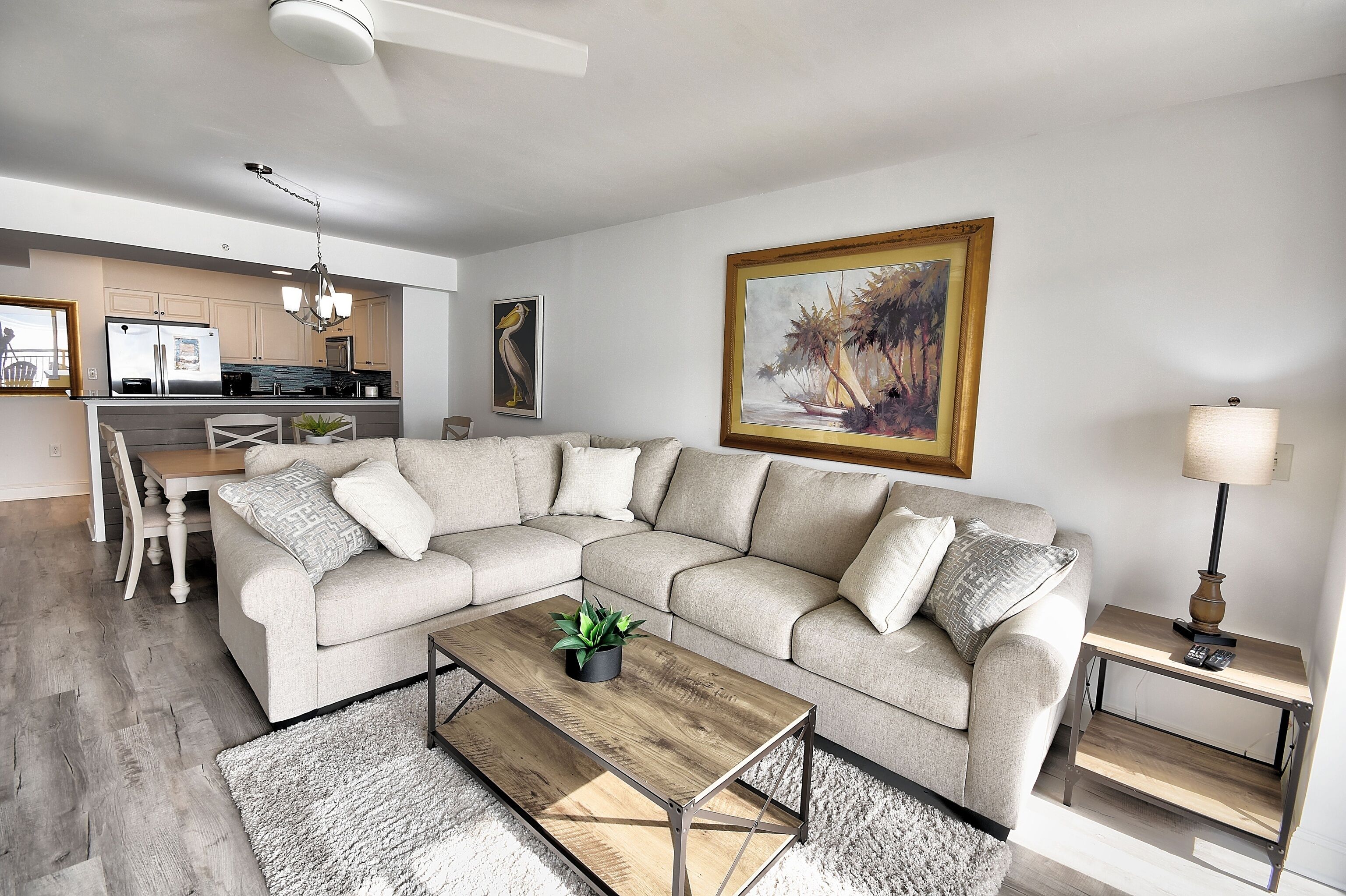A modern living room with a sectional sofa, a coffee table, a dining area with a chandelier, and a kitchen in the background.
