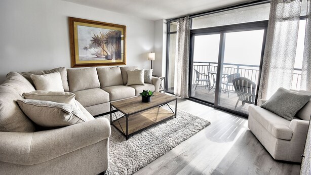 Ein Wohnzimmer mit einer Couch, einem Sessel, einem Couchtisch und einem großen Fenster mit Blick ins Freie.
