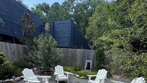 Een terras met Adirondack-stoelen rond een vuurkorf, omgeven door een houten hek en groen.