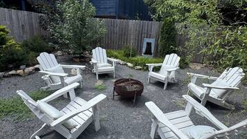 A deck with Adirondack chairs arranged around a fire pit, surrounded by a wooden fence and greenery.