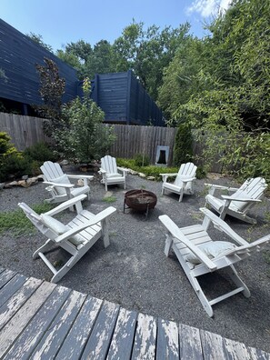 A deck with Adirondack chairs arranged around a fire pit, surrounded by a wooden fence and greenery.