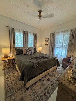 Une chambre à coucher comprenant un lit, deux tables de chevet avec des lampes, un ventilateur de plafond et une fenêtre avec des rideaux.