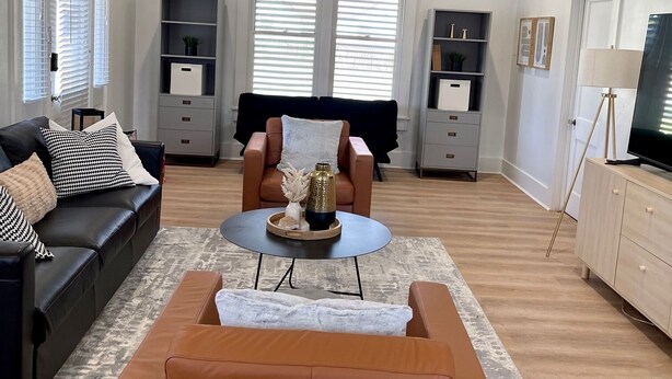 A living room with a ceiling fan, a black sofa, a brown leather couch, a coffee table, and a TV on a wooden cabinet. - Gem on Spring-Upscale home walk to Main sleeps 11 (Del Rio)