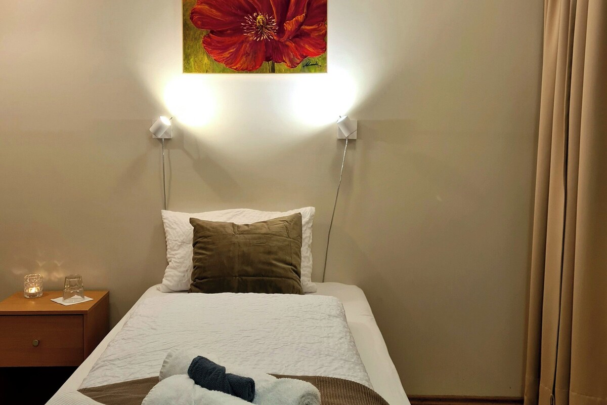Hotelkamer met een bed, nachtkastje, schilderij van een rode bloem en raam met gordijnen.