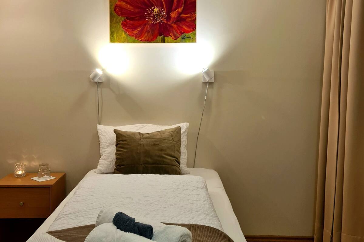 Una habitación de hotel con cama, mesita de noche, un cuadro de una flor roja y una ventana con cortinas.
