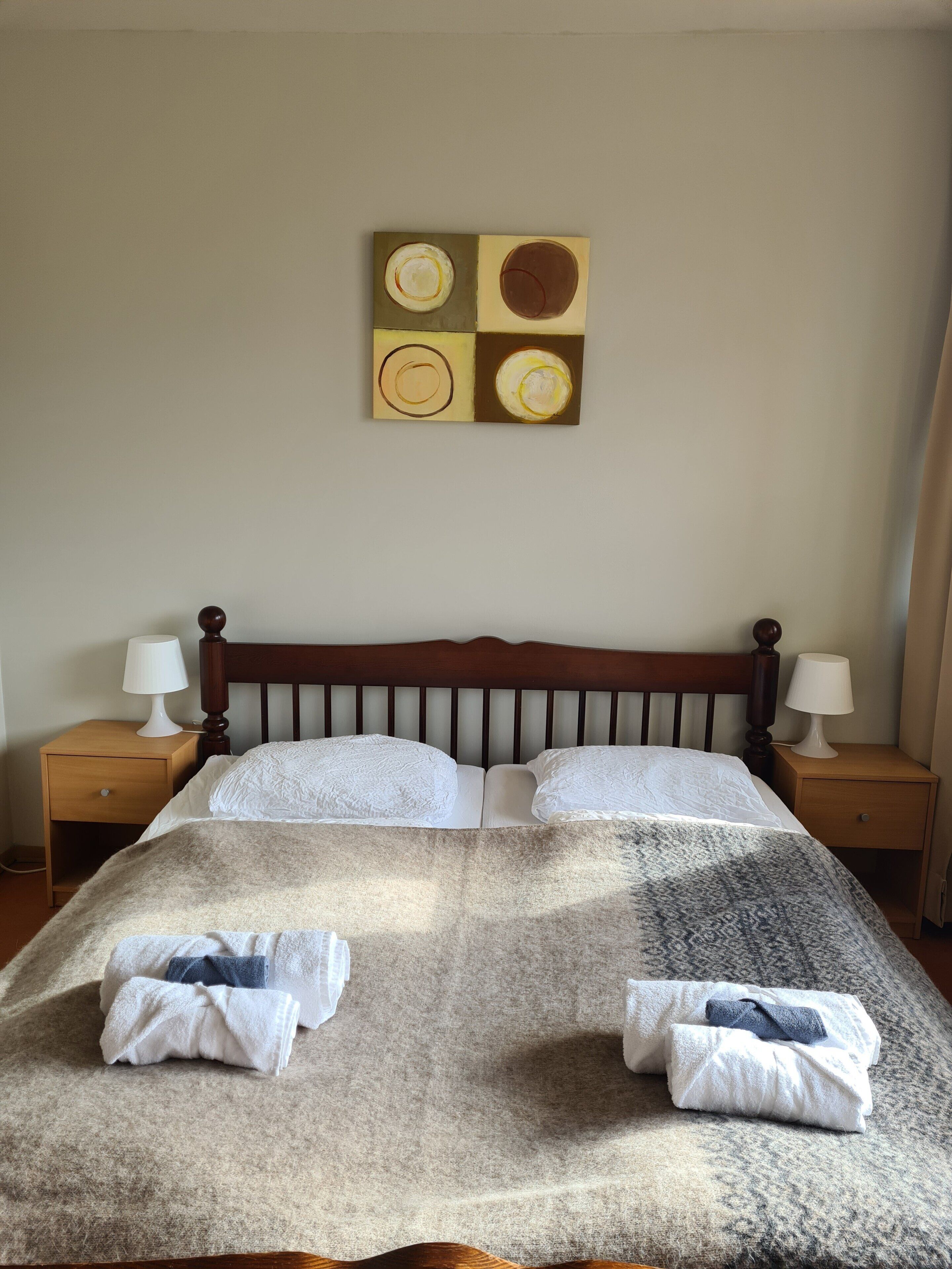 Una cama con cabecera, dos mesitas de noche con lámparas, y una obra de arte en la pared sobre la cama.