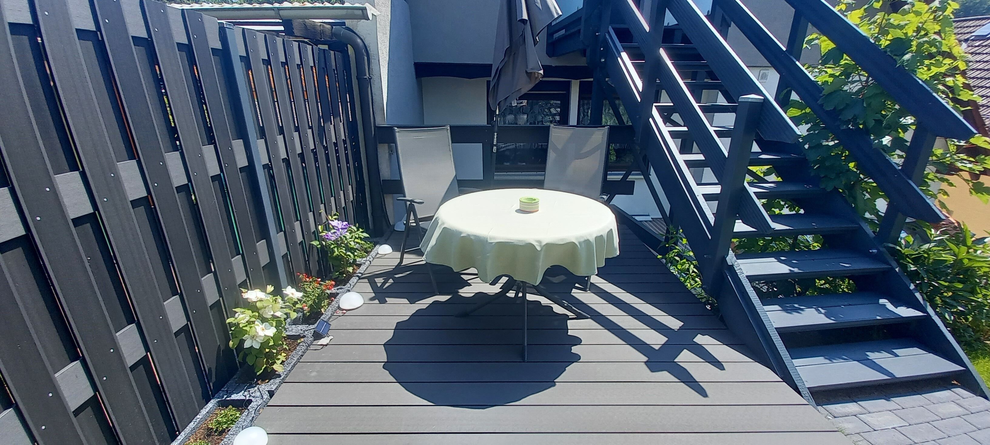 Een terras met een ronde tafel en stoelen, een houten hek en een trap.