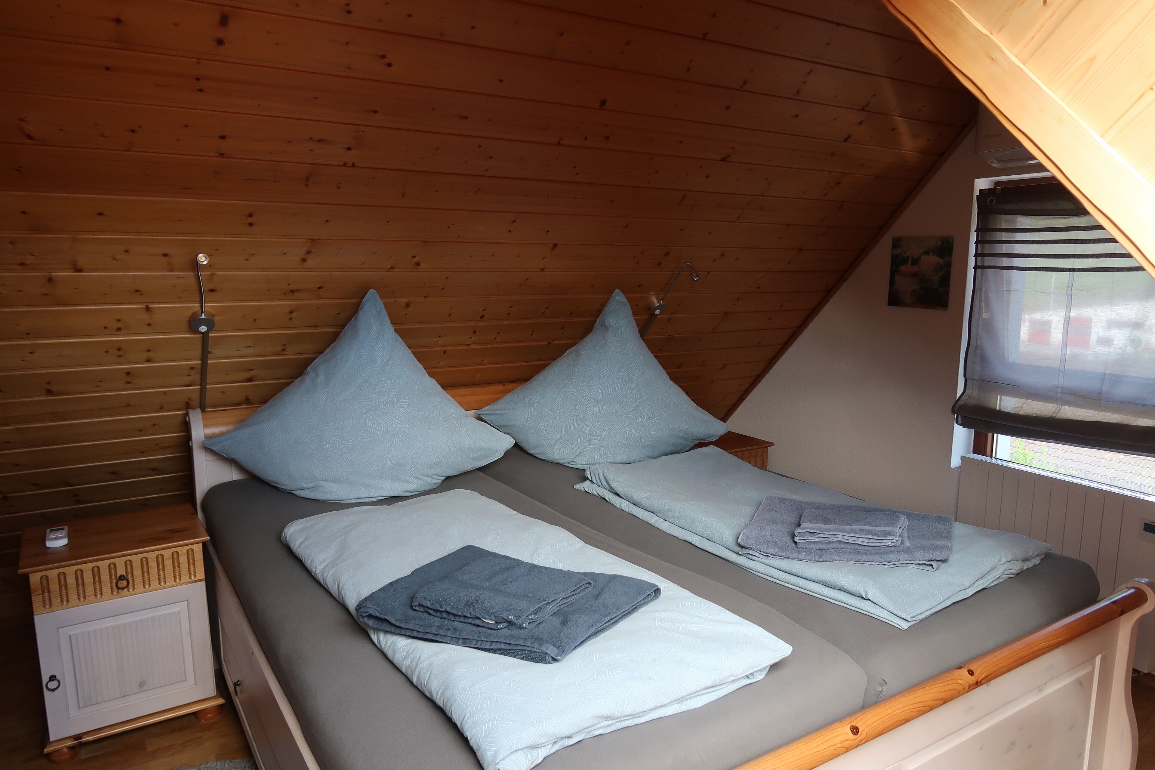 A small, cozy room with a sloped ceiling and wooden walls. It features two single beds with matching pillows and folded towels.