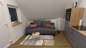 Un espacio habitable compacto con un sofá gris, una mesita de centro redonda, una cocina con armarios grises, una encimera de madera y una ventana con vistas.