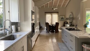A modern kitchen with white cabinetry, a central island, and a dining area with chairs. - Welcome to your island home, Mystical Mermaid! (Cruz Bay)