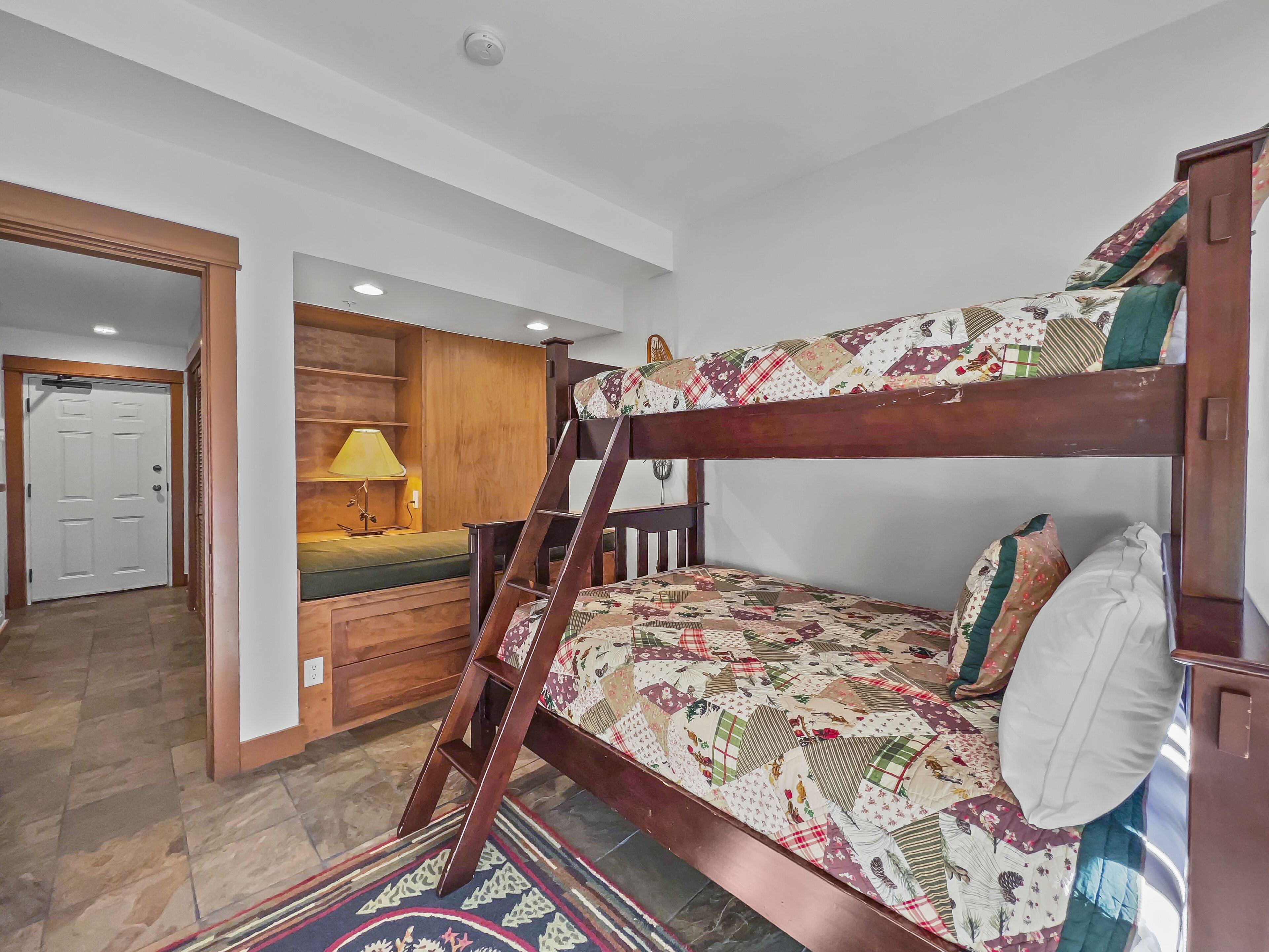 A bunk bed with a quilted cover, a wooden desk with a lamp, and a door in the background.