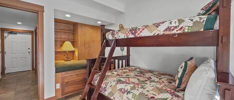 A bunk bed with a quilted cover, a wooden desk with a lamp, and a door in the background.