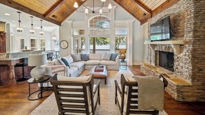 A spacious living room with a high ceiling, a stone fireplace, and a large window overlooking the outdoors.