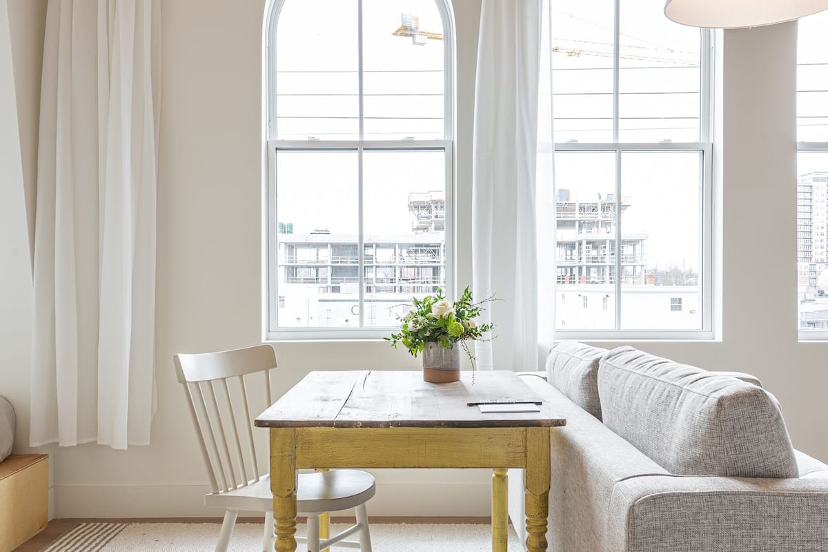 Una habitación con techo de madera, paredes blancas y ventanas arqueadas grandes. Hay una silla blanca, una mesa pequeña con una planta y un sofá gris.