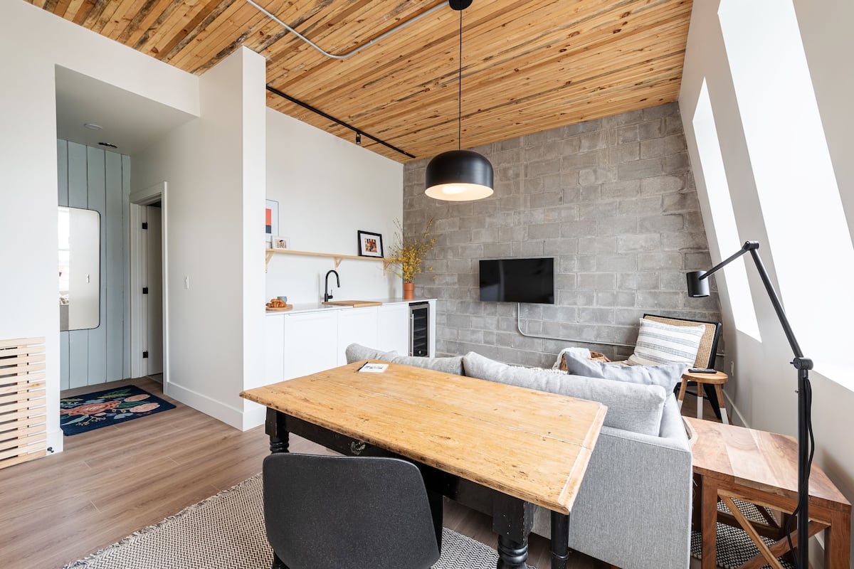 Un espacio habitable moderno con techo de madera, un sofá gris, una mesa de comedor de madera y un televisor de pantalla plana montado en una pared de ladrillo.