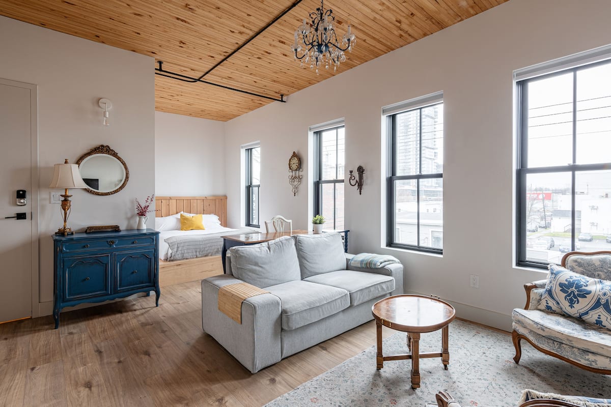Una sala moderna con techo de madera, cómoda azul, sofá gris, mesa redonda de madera y ventanas amplias que ofrecen vistas de la ciudad.