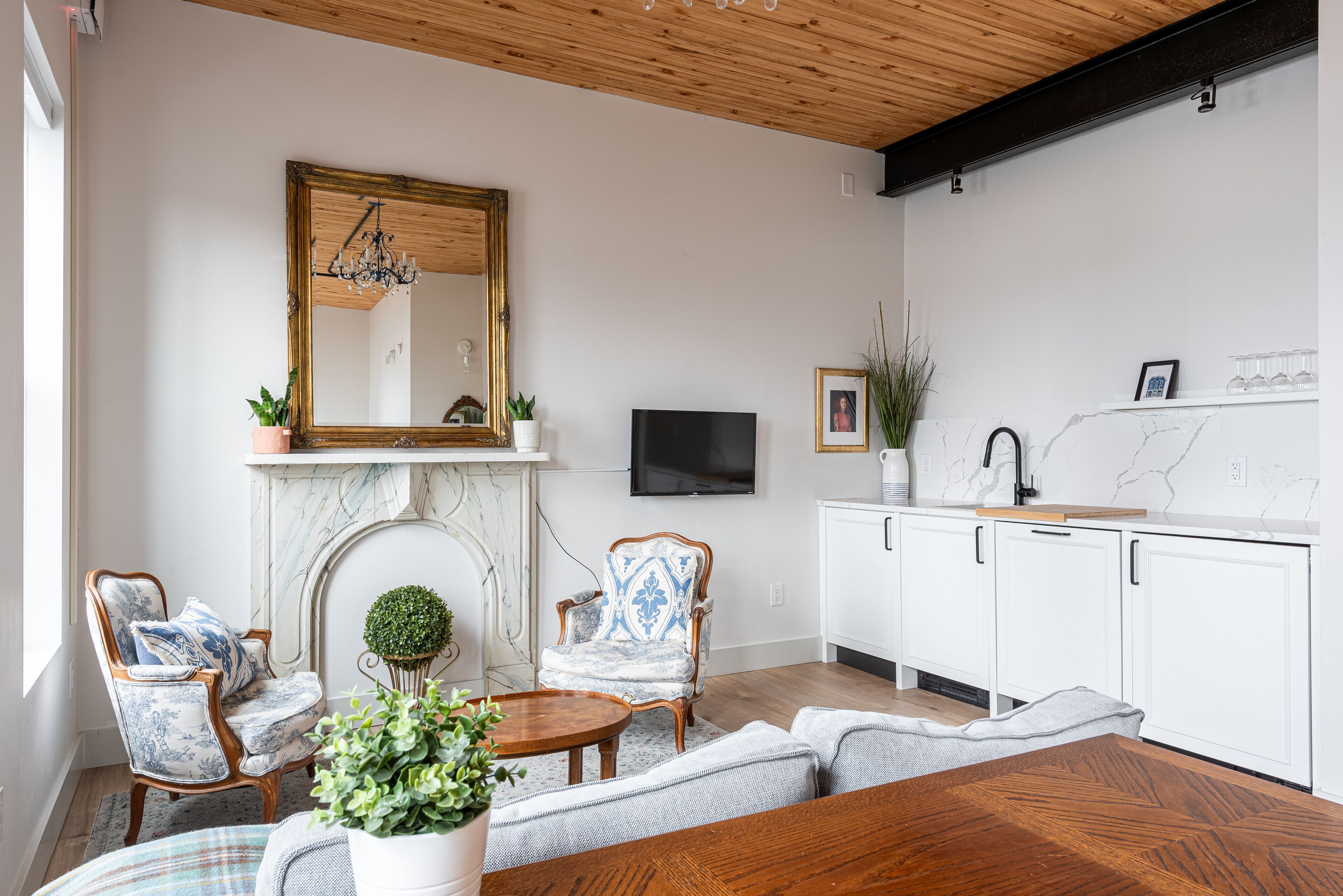 Ein modernes Wohnzimmer mit Kamin, großem Spiegel und Fernseher. Zwei Sessel, ein runder Tisch und ein Sofa sind ebenfalls vorhanden.