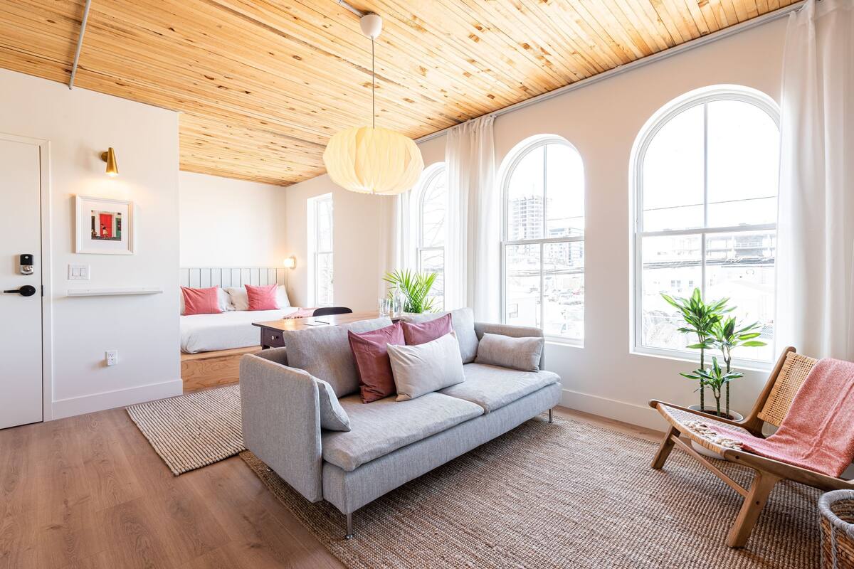 Un salón moderno con techo de madera, un sofá gris, una silla de madera y ventanas grandes.