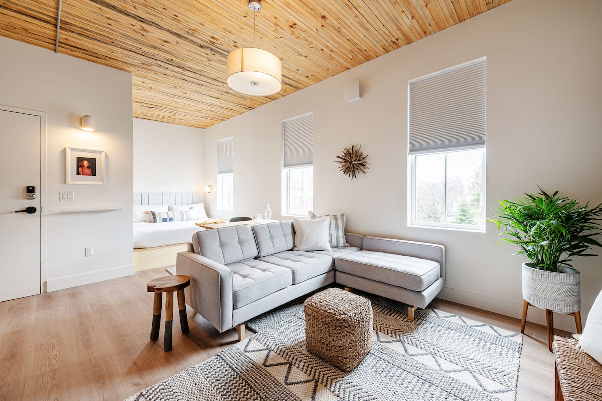 Una sala de estar moderna con un sofá seccional gris, techo de madera y un ventanal con persianas.