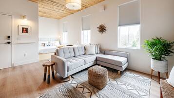 Un salon moderne avec un canapé d’angle gris, un plafond en bois et une grande fenêtre équipée de stores.