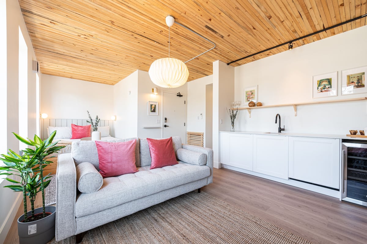 Una sala de estar moderna con un sofá gris, cojines rojos, techo de madera y una cocina con armarios blancos.