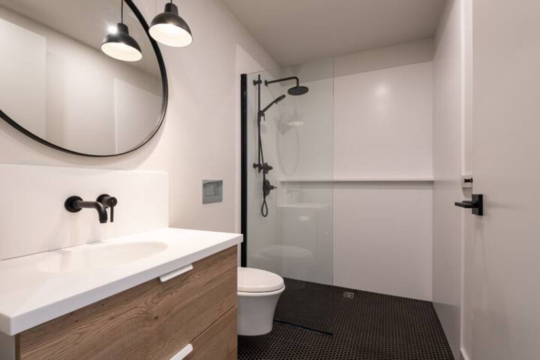 Un baño moderno con lavamanos blanco, tocador de madera y cabina de ducha con mampara de vidrio.