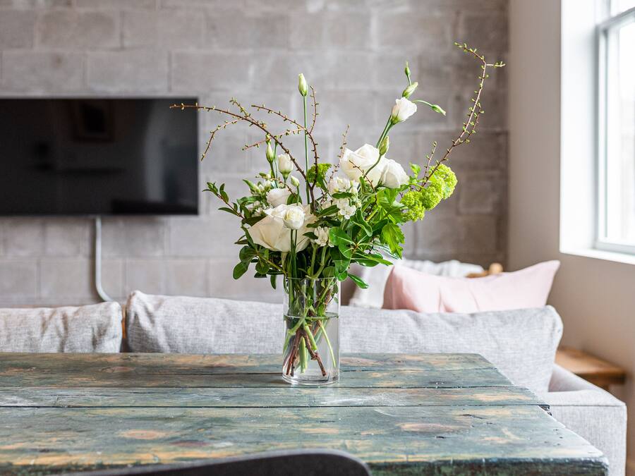 Una mesa de comedor rústica con un florero, un televisor montado en una pared de ladrillo, y una ventana con vistas.