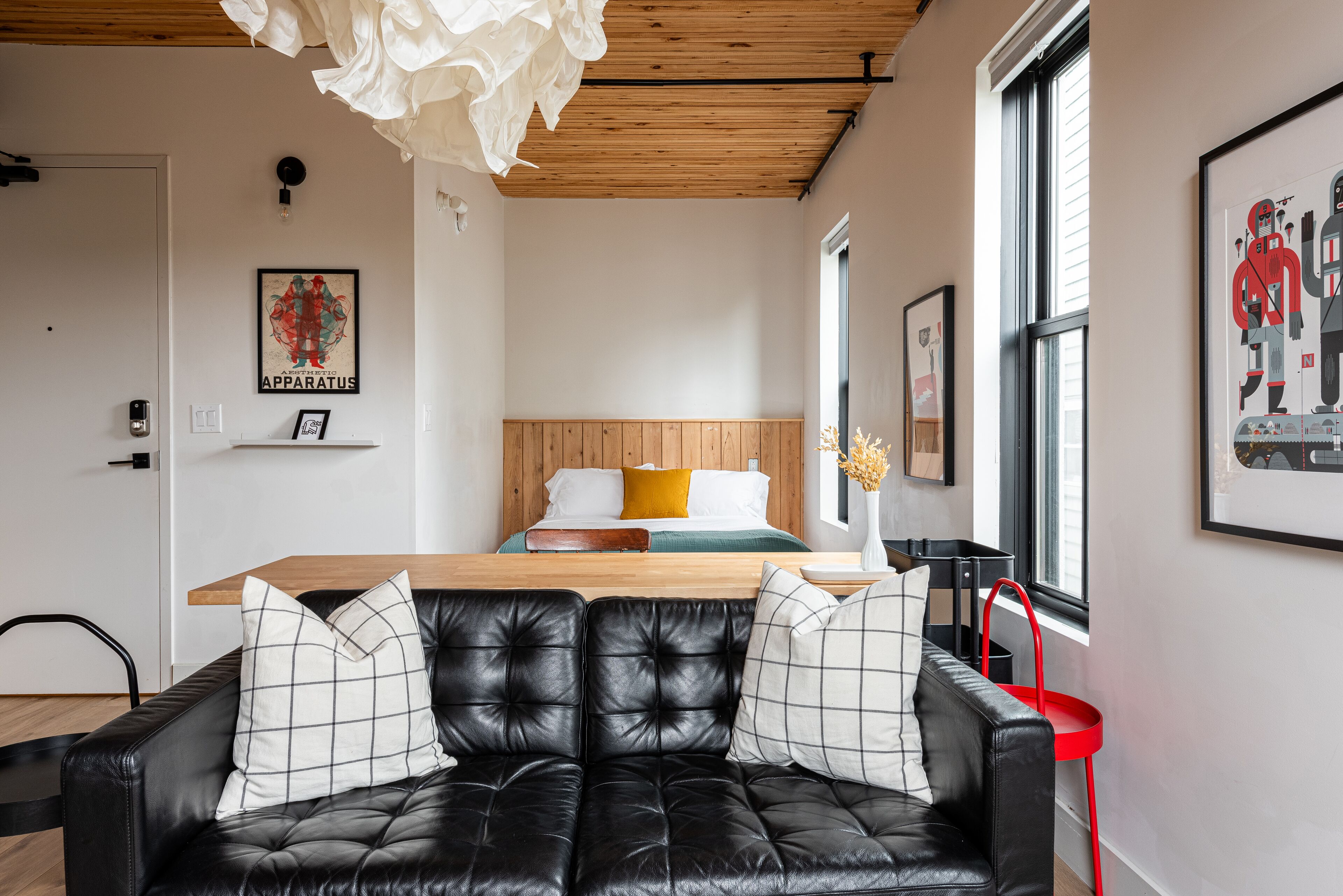 A modern living room with a black leather sofa, a wooden dining table, and a bed with a wooden headboard.