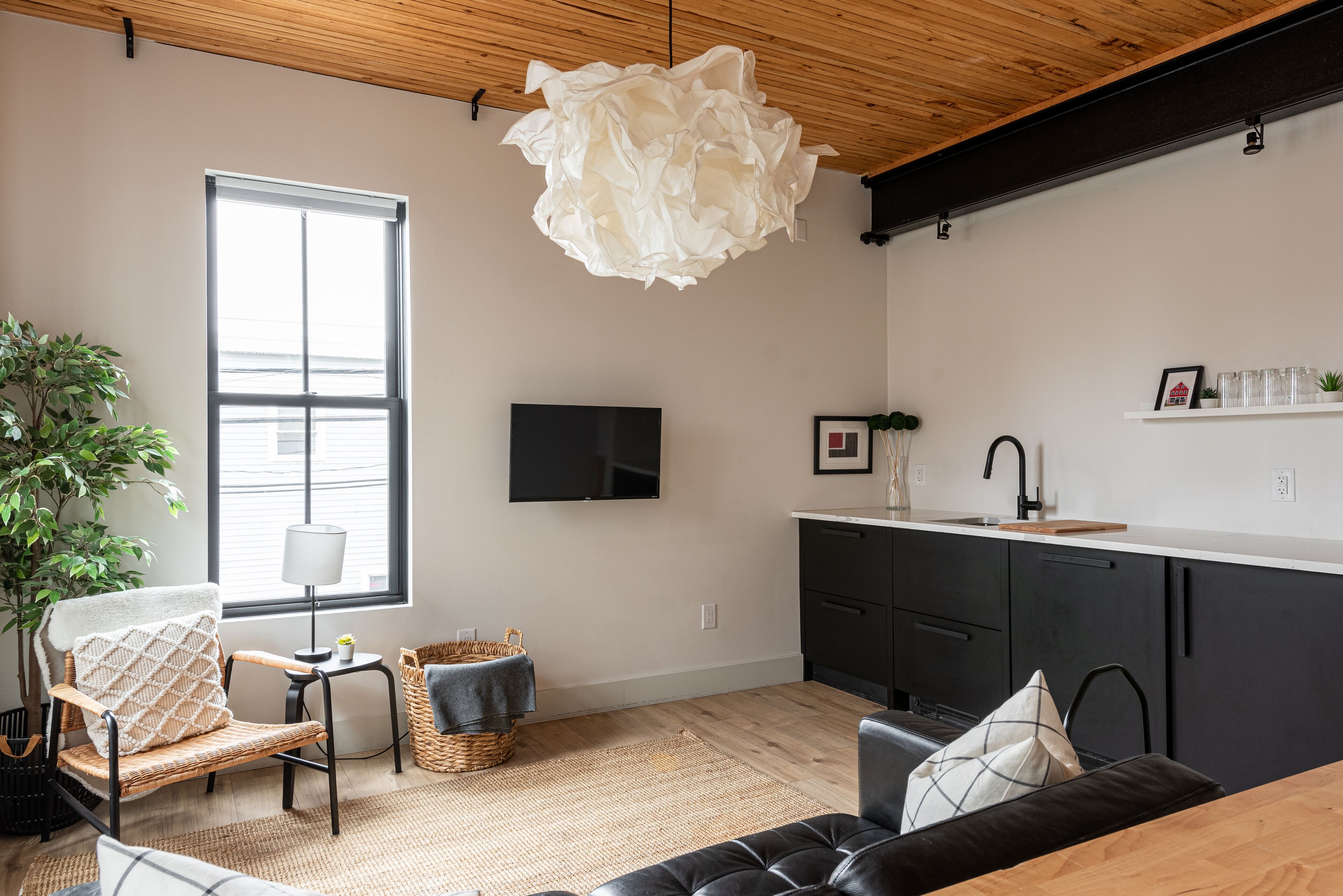 A modern living room with a large window, a flat-screen TV, a dining area with a kitchen in the background, and a cozy seating area.