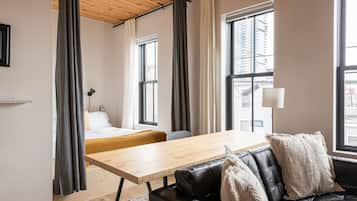 A modern room with a bed, a wooden table, a leather sofa, and large windows offering a city view.