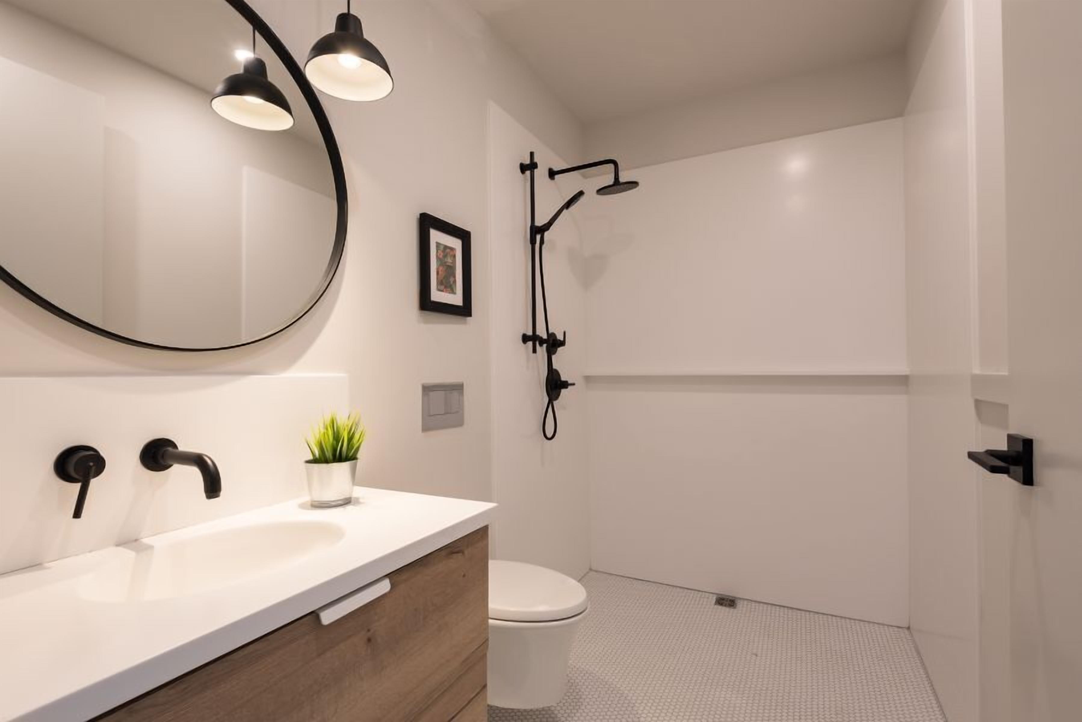 Un baño moderno con un lavamanos blanco, grifo negro y un espejo grande. Hay una pequeña planta en maceta sobre el mostrador y un cuadro en la pared.