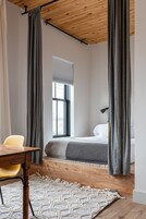 A bedroom with a wooden ceiling, a bed with a gray blanket, a bedside lamp, and a window with blinds.