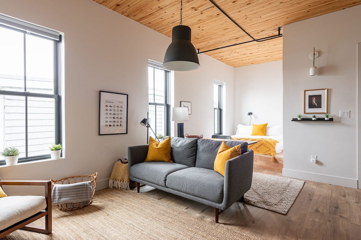 Una sala de estar moderna con un sofá gris, cojines amarillos, un sillón de mimbre y un ventanal grande.
