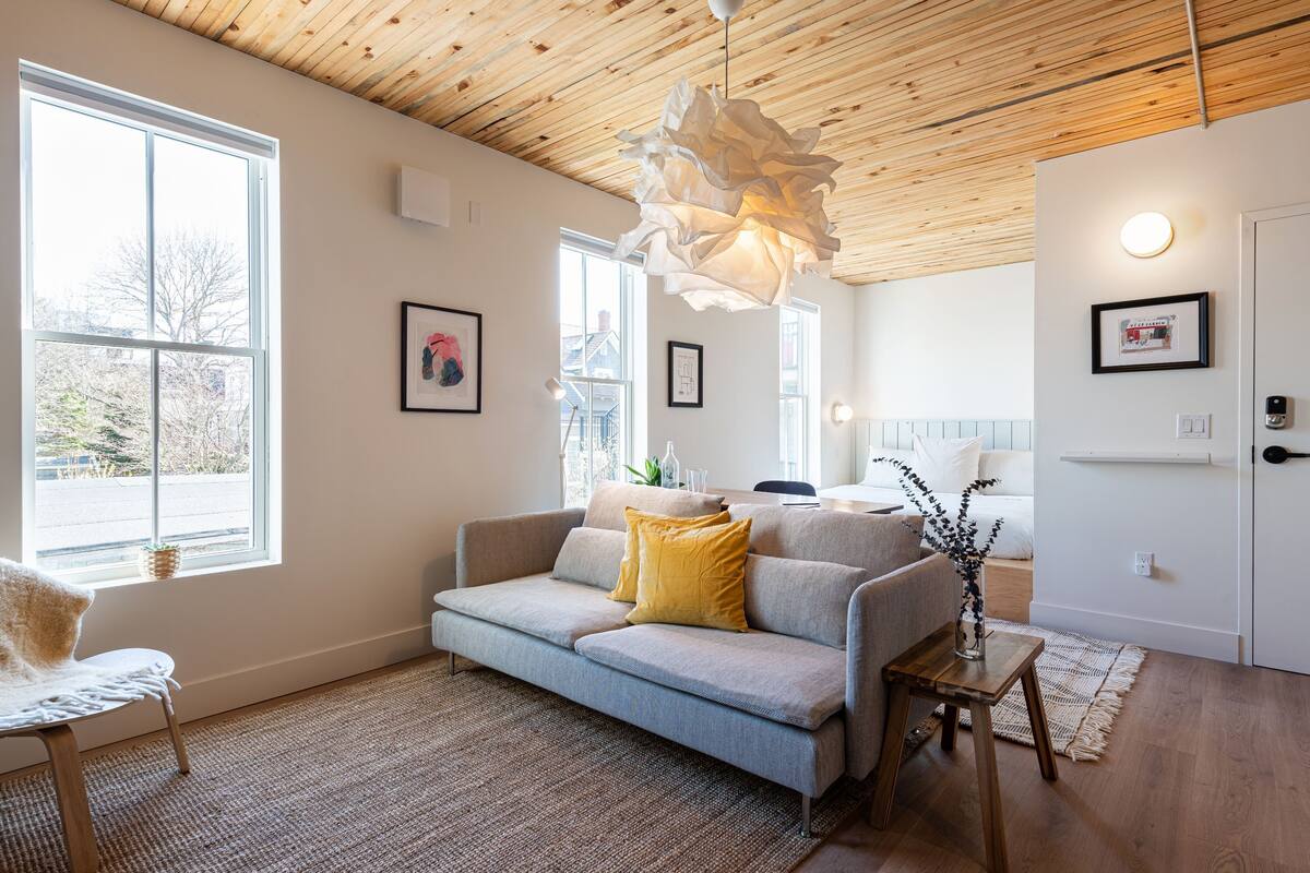 Una sala de estar moderna con un sofá gris, techo de madera y ventanas amplias.