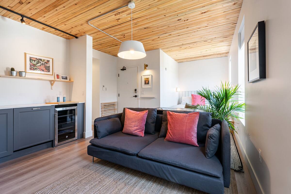 Un espacio de estar moderno con techo de madera, un sofá gris oscuro con cojines rosados, una zona de cocina con armarios grises y una planta grande.