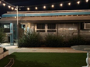 Un edificio moderno con techo plano, toldo azul y fachada de metal. Hay luces colgantes arriba y un pequeño jardín con césped y arbustos.