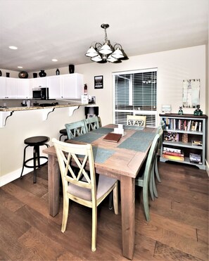A dining area with a wooden table and chairs, a kitchen with white cabinets, and a bookshelf with books.