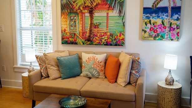 A living room with a beige sofa, patterned cushions, a glass coffee table, and a painting of a tropical scene. - Casa Louisa -Beautiful 2 Bedroom in Old Town - Right off Duval/Great Front Porch (Key West)