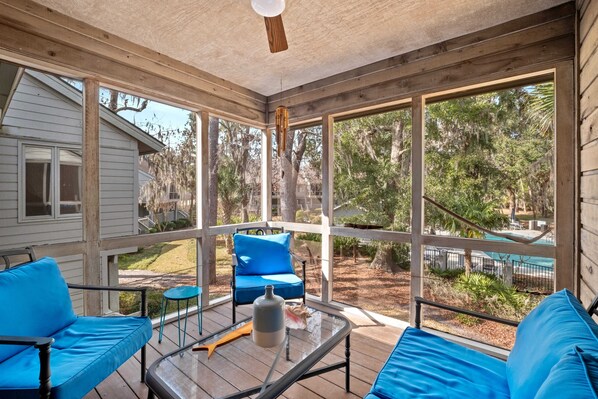 A covered porch with blue furniture, a glass table, and a view of a pool and trees. - Spacious condo enjoy Pool, Hot Tub access to Beach with Private Beach Locker ! (Hilton Head Island)