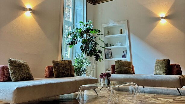 Ein Zimmer mit hoher Decke, verziert mit aufwendigen Stuckarbeiten und einem großen Fenster. Zwei Sofas mit Kissen sowie ein Glastisch sind ebenfalls vorhanden.
