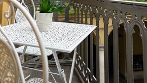 Ein weißer Metalltisch mit einem Blumentopf, platziert auf einem Balkon mit aufwendiger Metallgeländer und zwei großen Fenstern im Hintergrund.