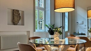 Ein Essbereich mit einem Glastisch, umgeben von Stühlen, einem großen Fenster mit Blick auf Grünflächen und einem Heizkörper an der Wand.