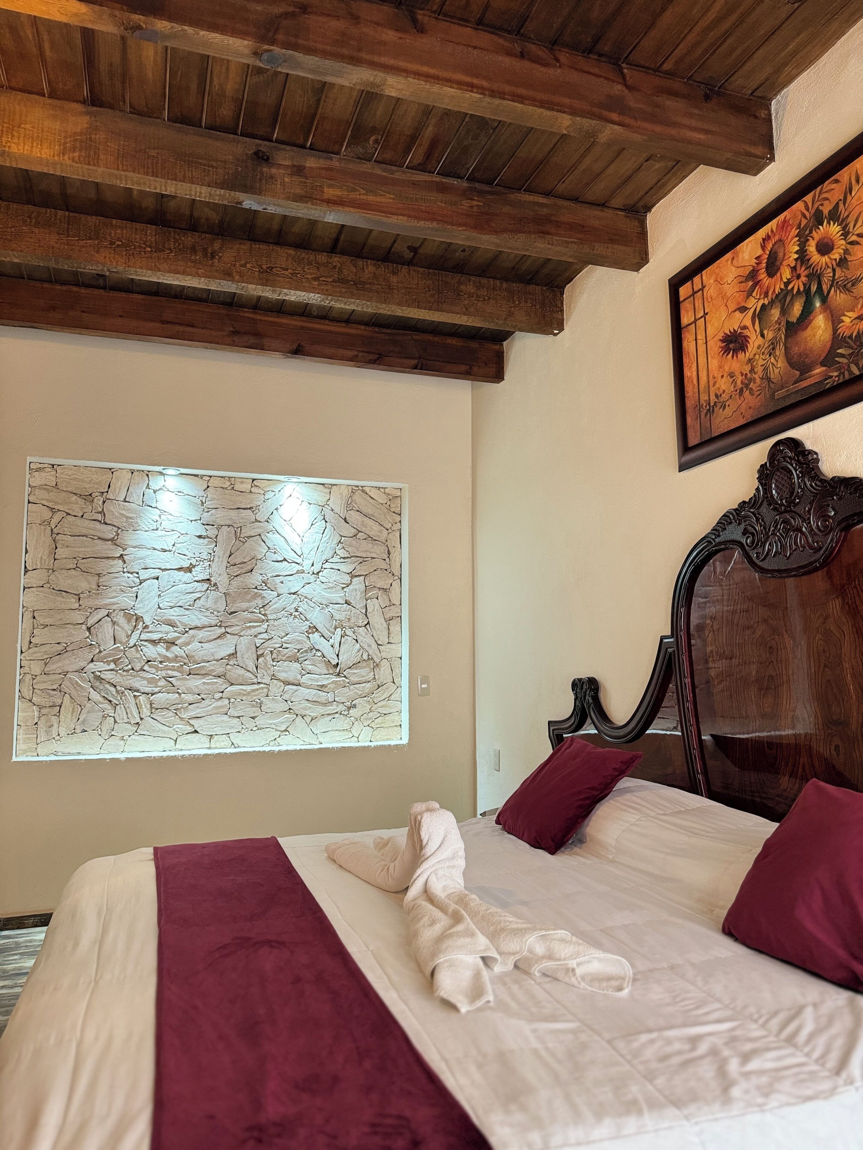 A neatly made bed with a white and maroon bedspread, a stone wall art piece, and a wooden ceiling.